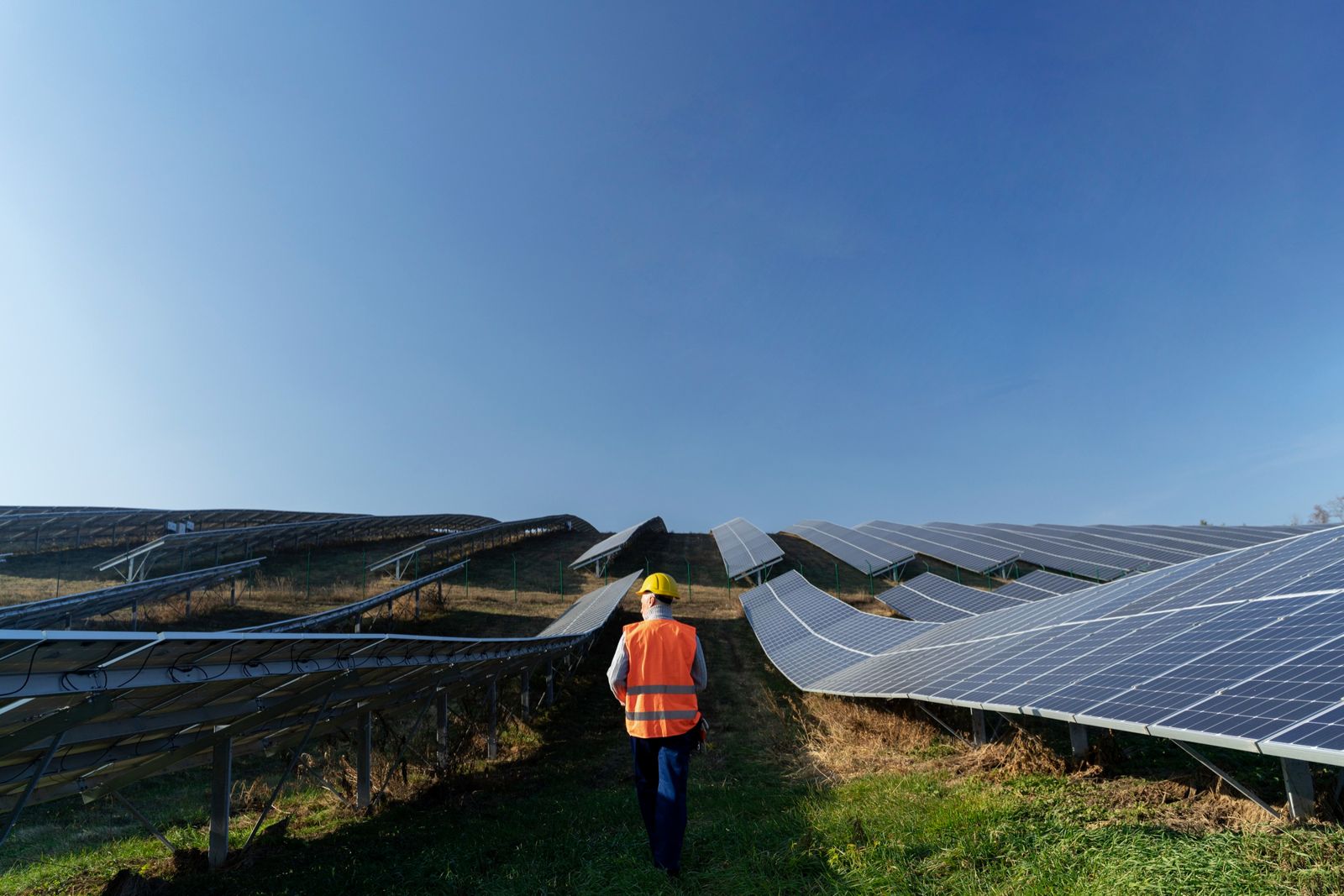 Técnico de capacete e colete laranja caminhando entre as fileiras de placas de uma usina solar